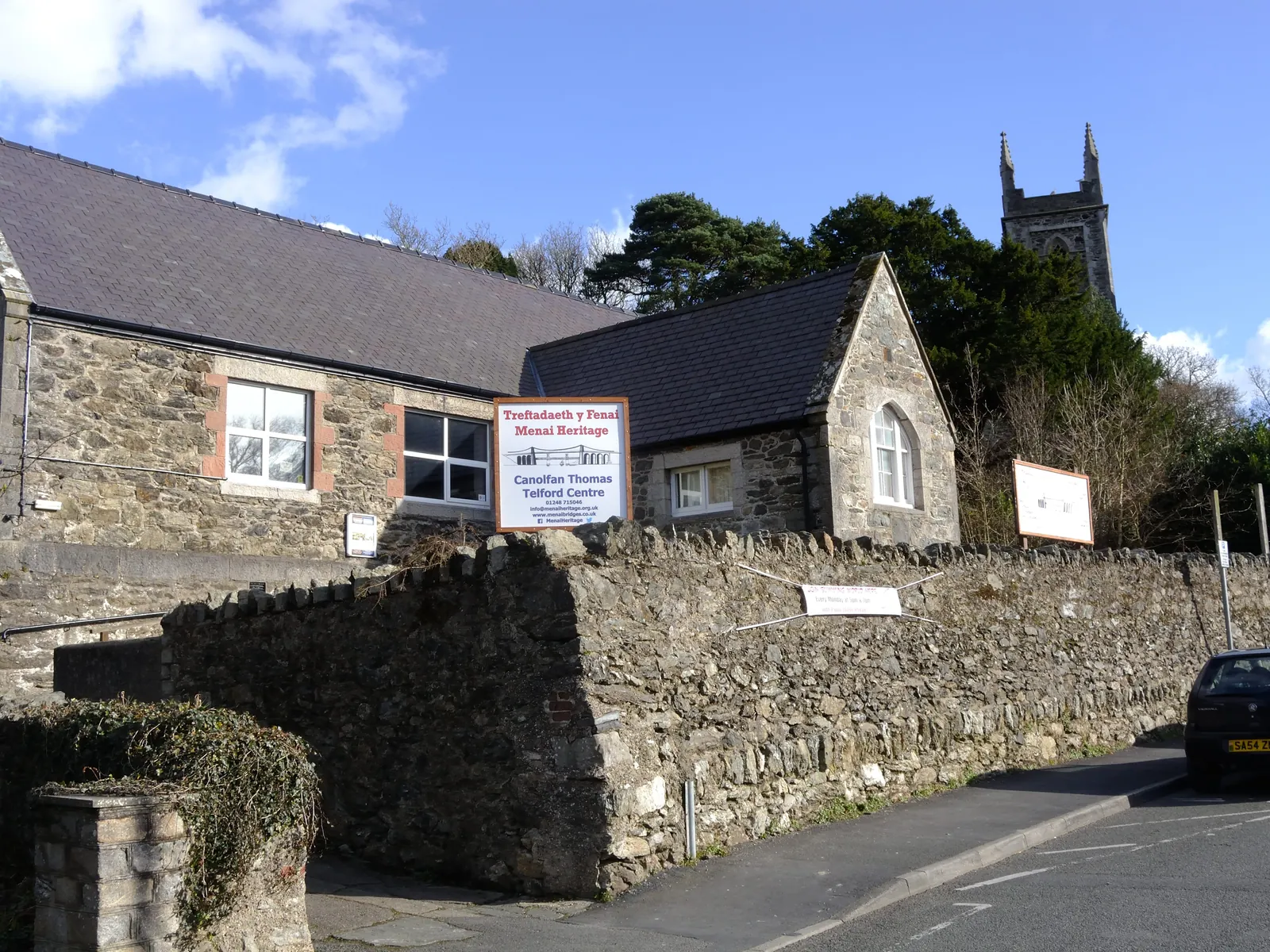 The Thomas Telford Centre on Mona Road, Menai Bridge: a stone former school building carrying the Menai Heritage sign, a church tower behind