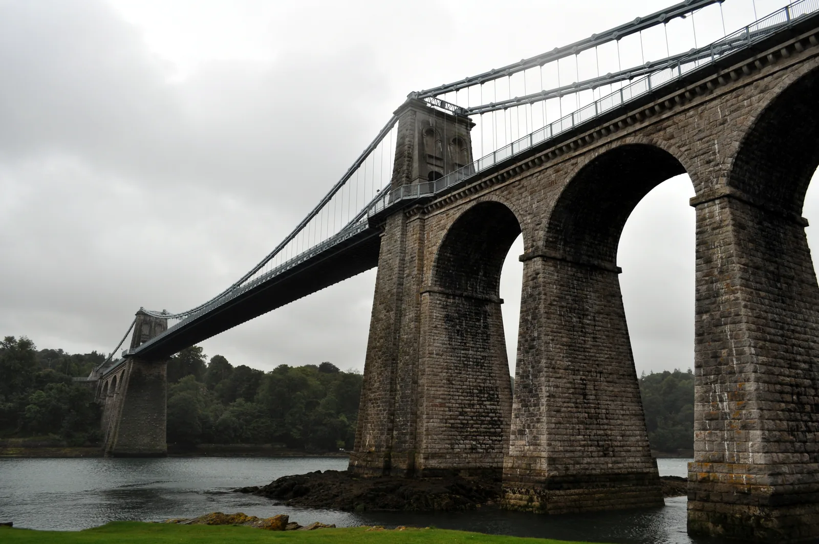 The stone approach arches of the Menai Suspension Bridge standing in the water of the strait, the suspended span carrying away to the left
