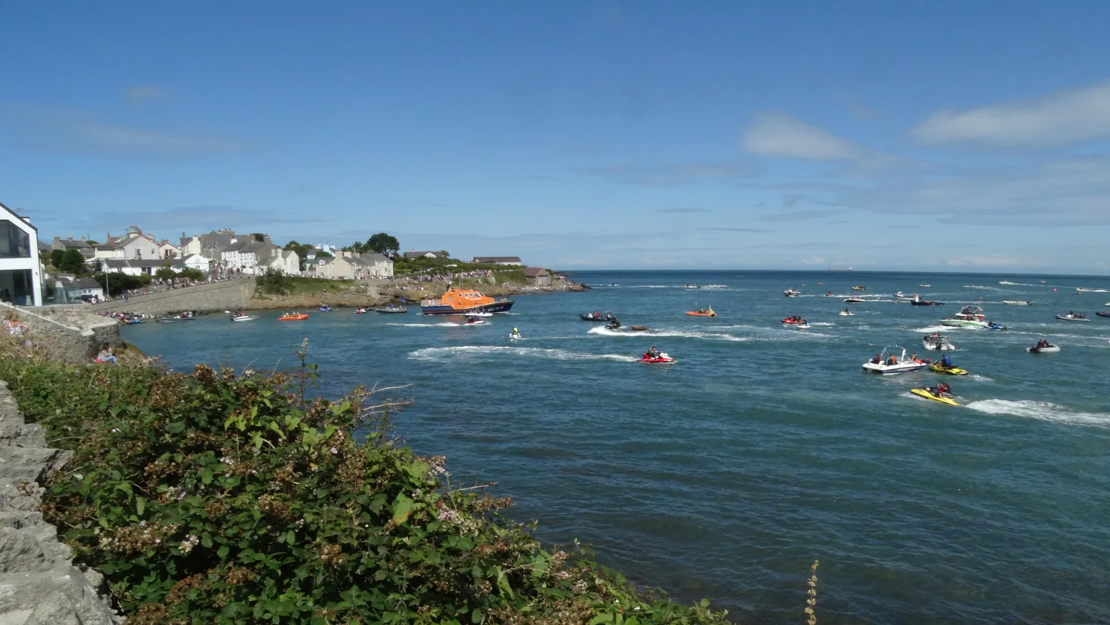 Small boats and inflatables out on the water in Porth Moelfre on Lifeboat Day, the village and its slipway behind