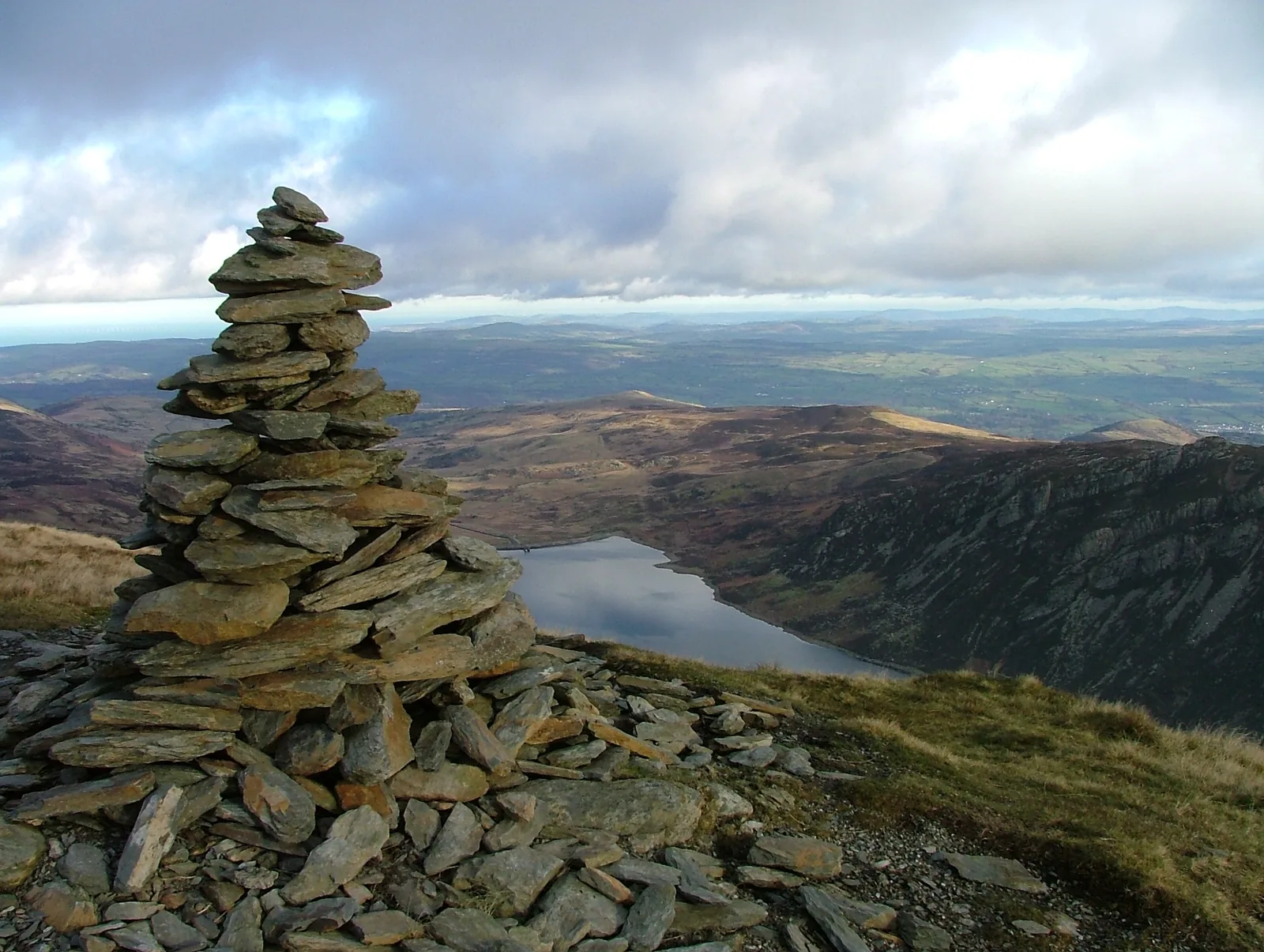 The summit cairn of Pen Llithrig y Wrach in the foreground, with Llyn Cowlyd and its crag-lined eastern shore below and the coastal plain beyond