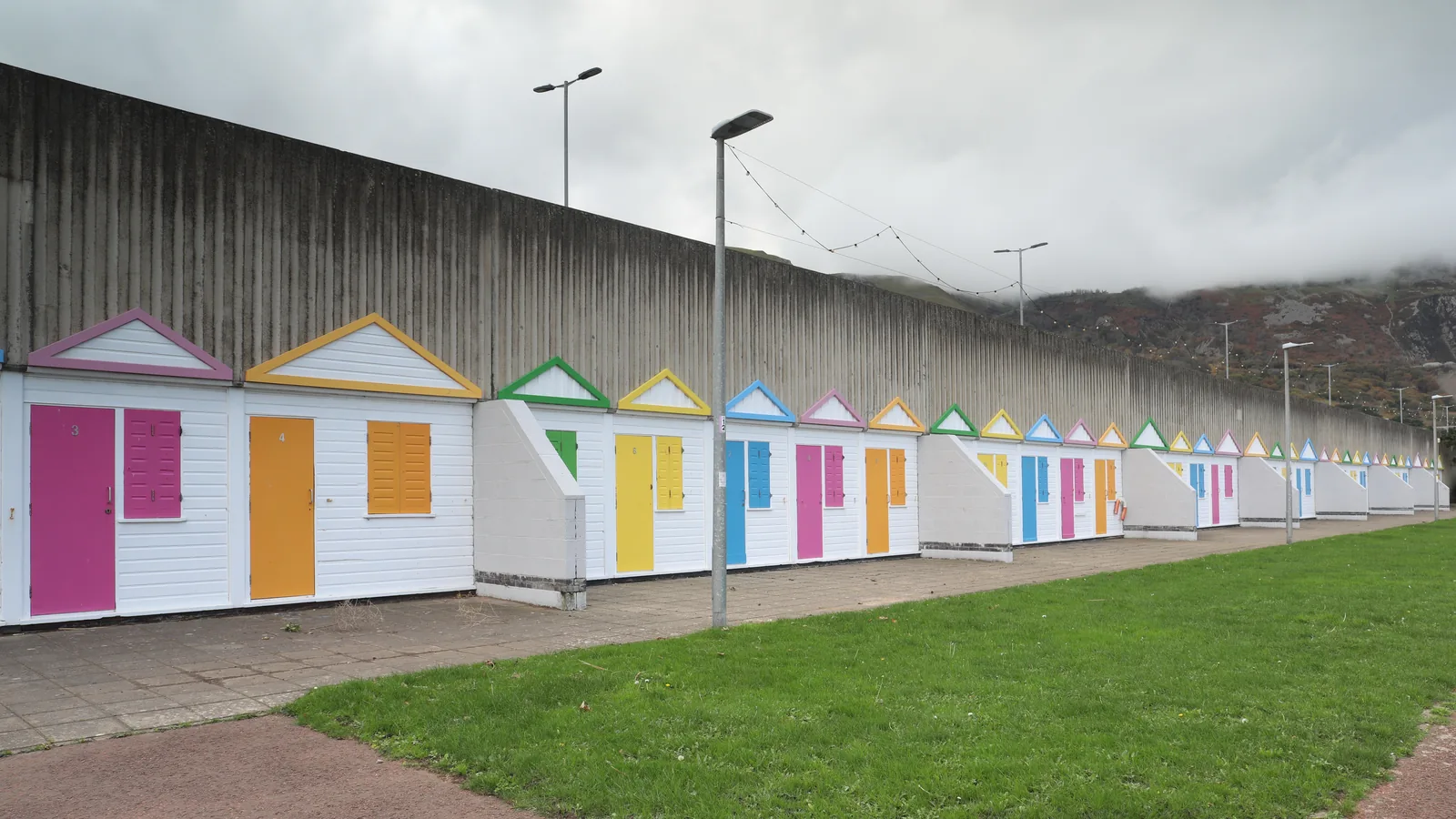 A row of painted beach huts along Penmaenmawr promenade beneath the tall retaining wall of the expressway
