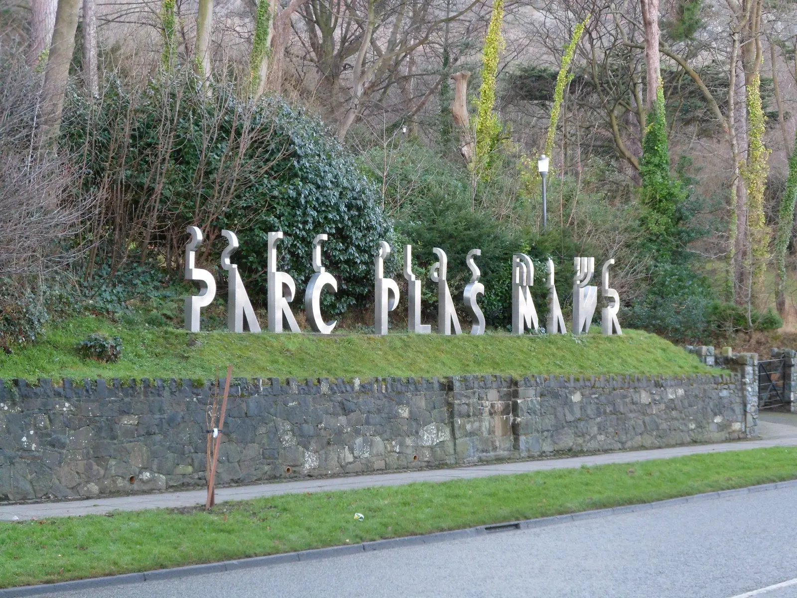 The name sculpture at the entrance to Parc Plas Mawr, Penmaenmawr, with the wooded slope of the park behind