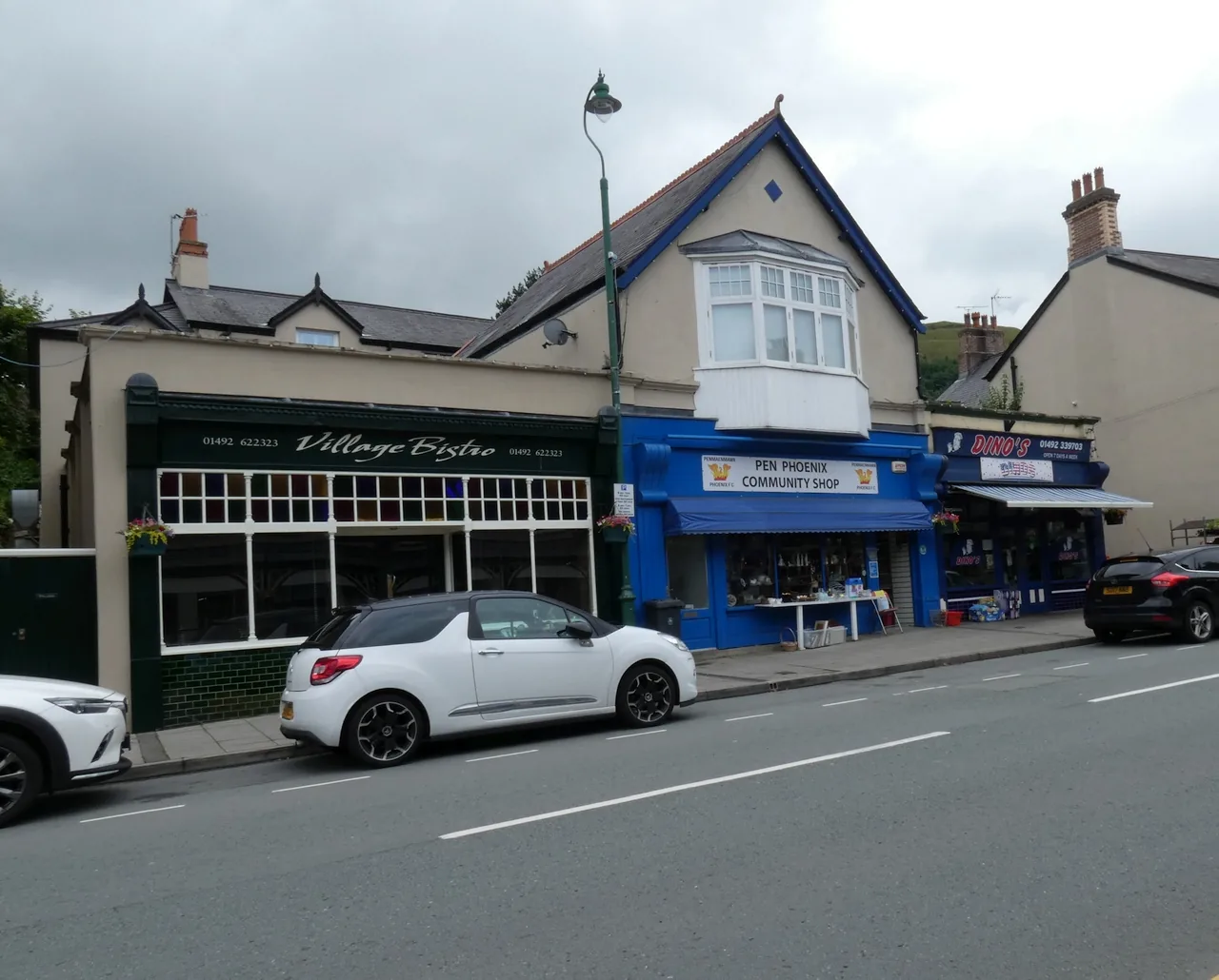 Independent shopfronts along Old Conway Road in Penmaenmawr, painted green and blue with cars parked outside