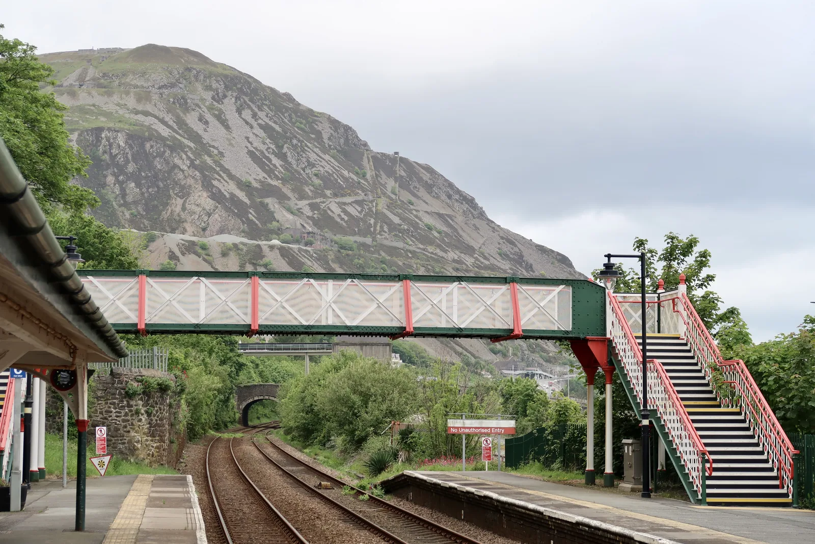 The Victorian footbridge at Penmaenmawr station, with the terraced grey face of the quarried mountain rising directly behind it