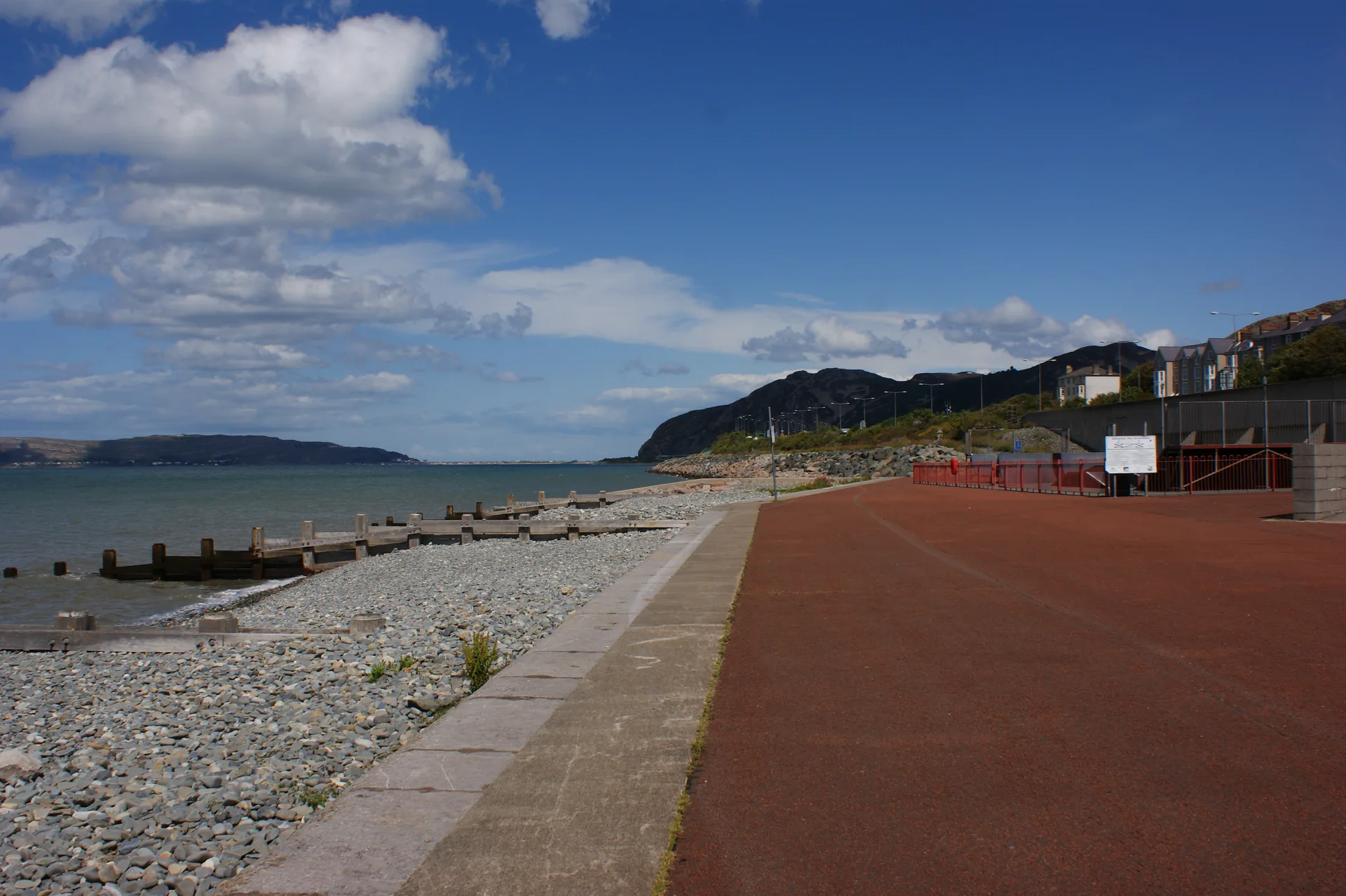 Penmaenmawr promenade running east beside shingle and timber groynes, with the headland of Penmaen-bach closing the bay and the town's terraces on the slope above