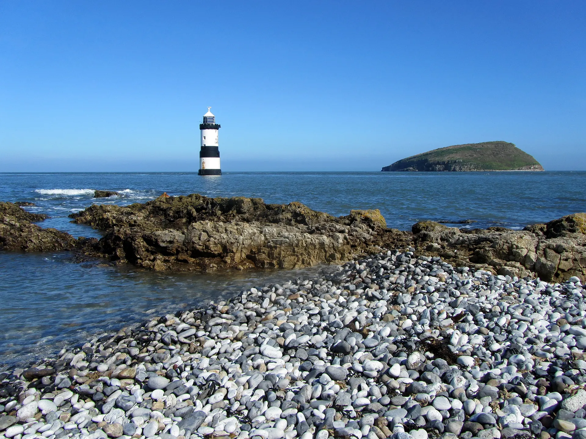 A wide view from Penmon Point across the sound, the black-and-white Trwyn Du Lighthouse offshore and Puffin Island low beyond.