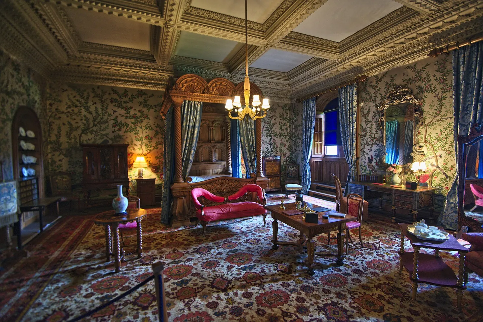 A grand carved four-poster bed and sitting area in one of Penrhyn Castle's state bedrooms, with patterned wallpaper and a chandelier.