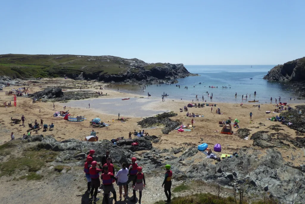 Porth Dafarch on a busy summer afternoon, groups sitting on the rocks above a beach of families, windbreaks and swimmers.