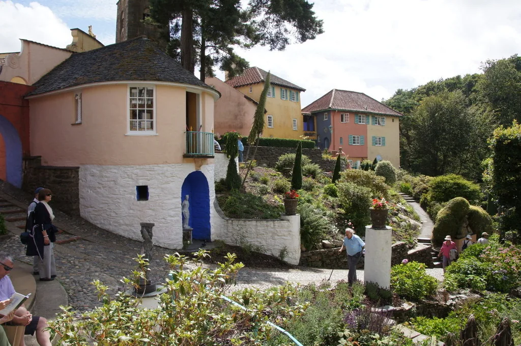A round tower building beside a sloping garden path with statues and topiary at Portmeirion, visitors walking through.