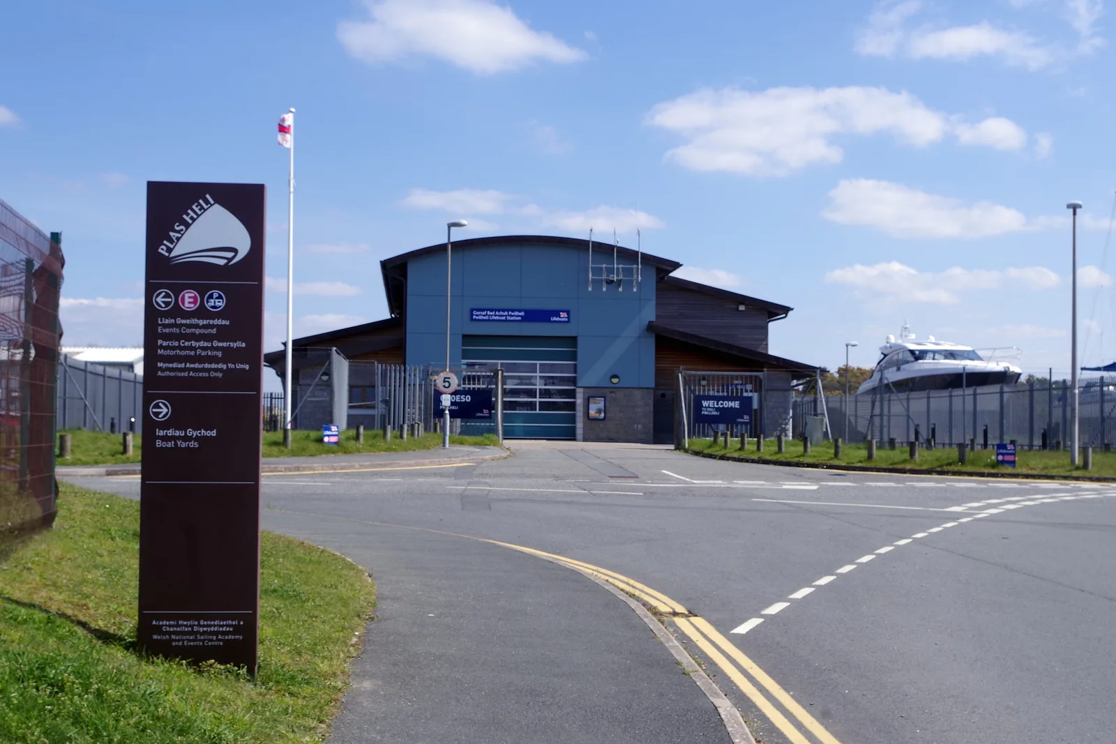 Pwllheli lifeboat station, a blue-clad boathouse behind its approach road, with a Plas Heli signpost in front