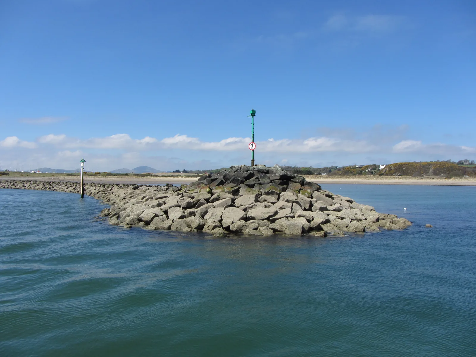 The rock breakwater and green navigation beacon at the entrance to Pwllheli harbour, sand and low hills beyond