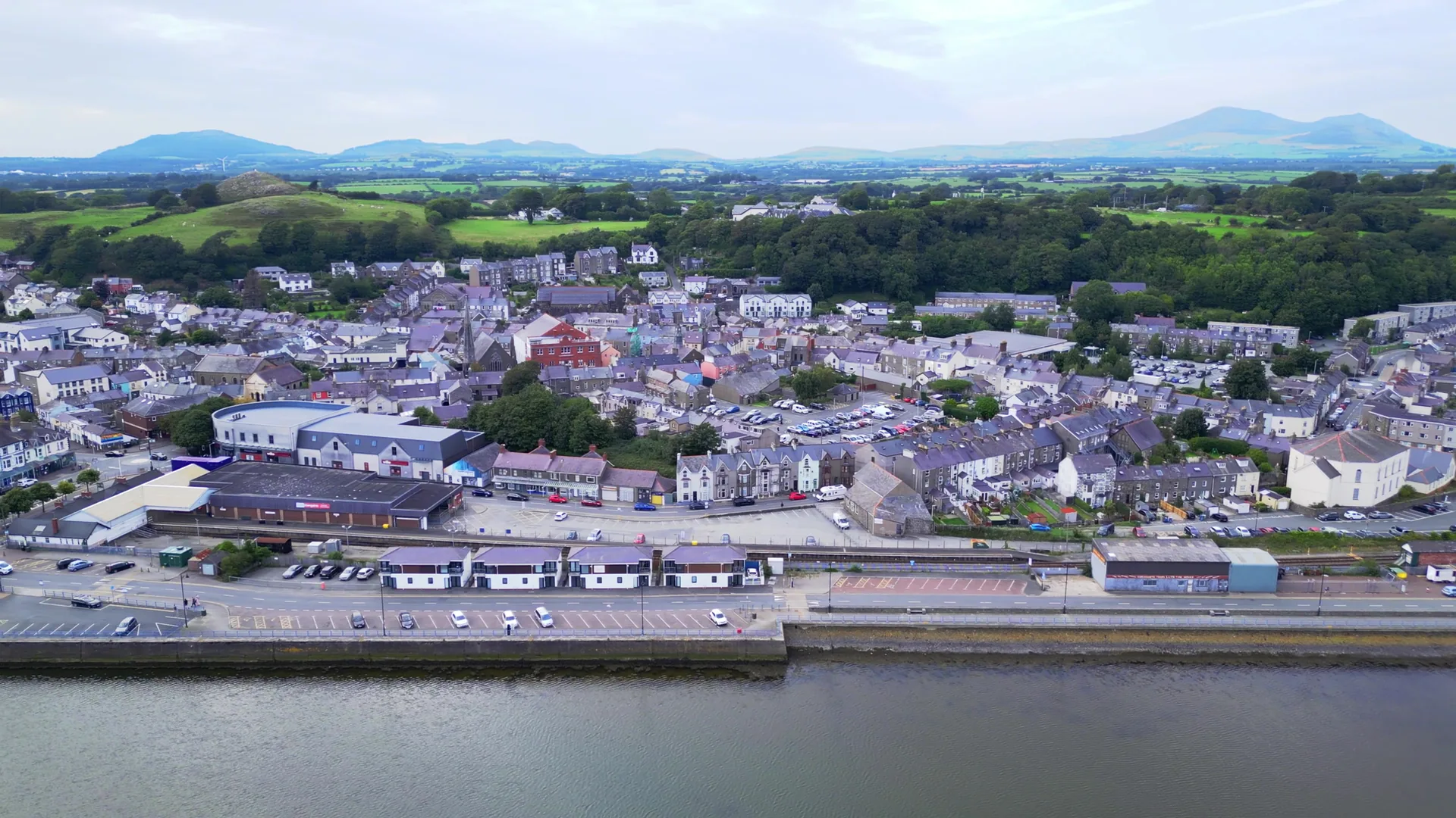 Pwllheli from the air above the Cob: the town&rsquo;s roofs and church spire behind the harbour wall, with the hills of the Llŷn on the horizon