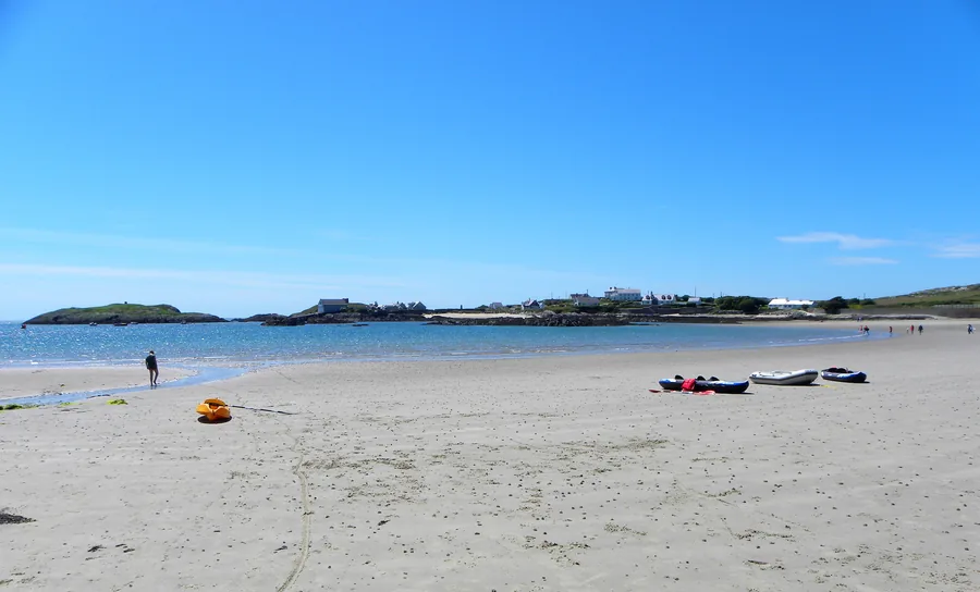 Borth Wen at Rhoscolyn: a wide sweep of sand above a sheltered blue bay, with a rocky islet and the old lifeboat house beyond.