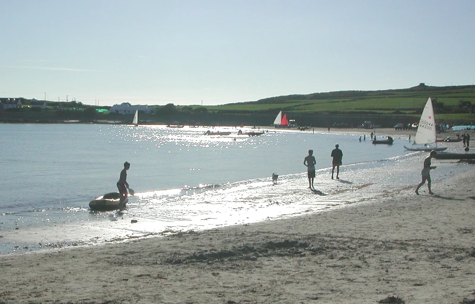People wading in the shallows at Borth Wen with sailing dinghies drawn up on the sand behind them and the headland beyond.