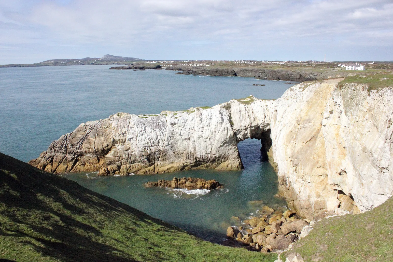 Bwa Gwyn, the White Arch, a pale rock arch cut through the headland cliff at Rhoscolyn with clear water beneath it.