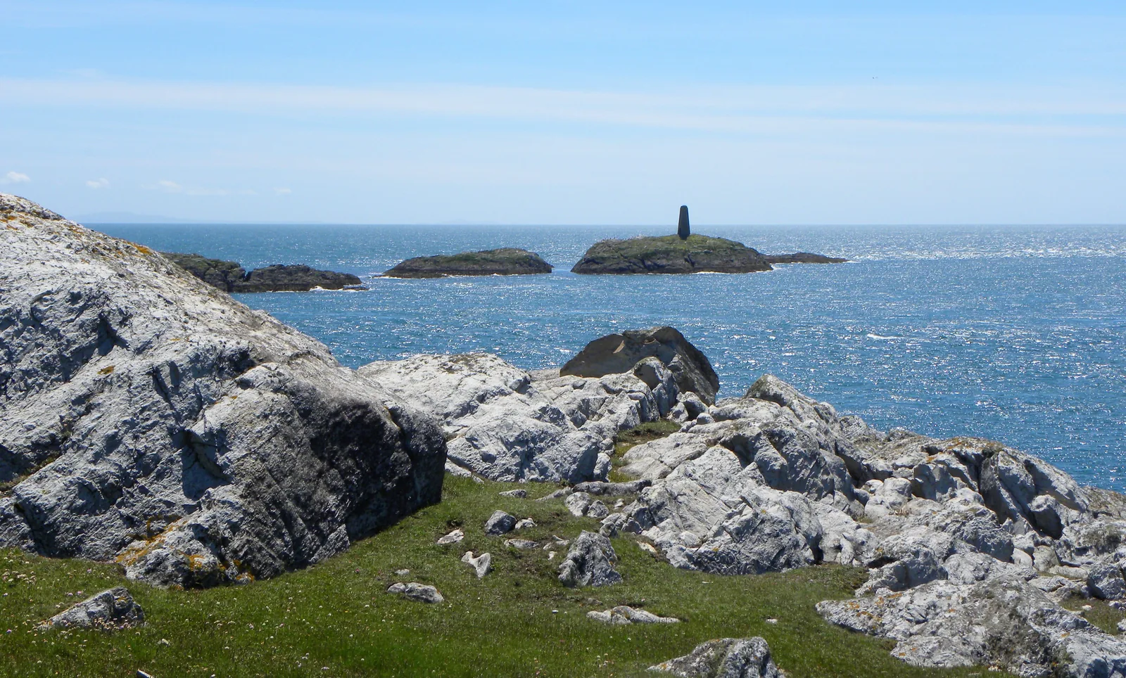 Rhoscolyn Beacon, a tall dark navigation marker standing on Ynysoedd Gwylanod offshore, seen across the water from the quartzite headland.