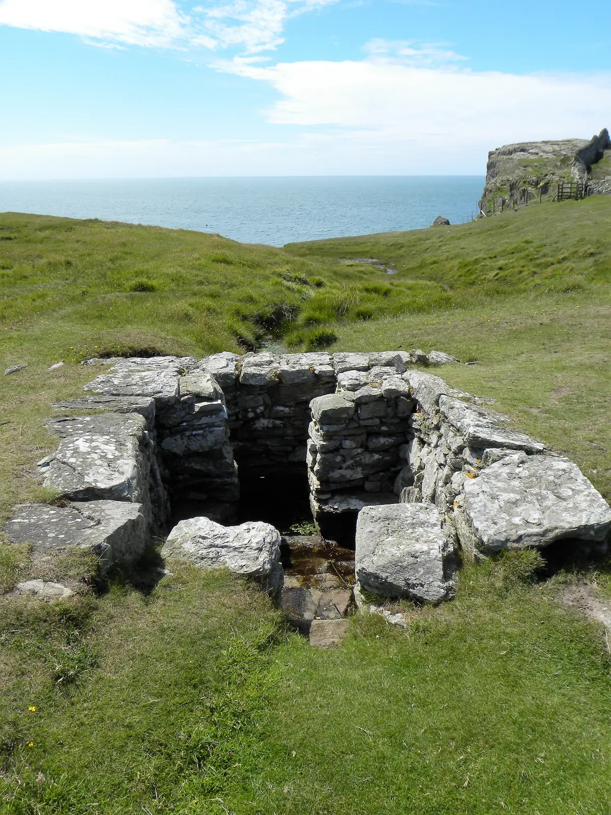 St Gwenfaen's Well at Rhoscolyn: a drystone well house sunk below the surrounding turf in a grassy dell, with the sea beyond.