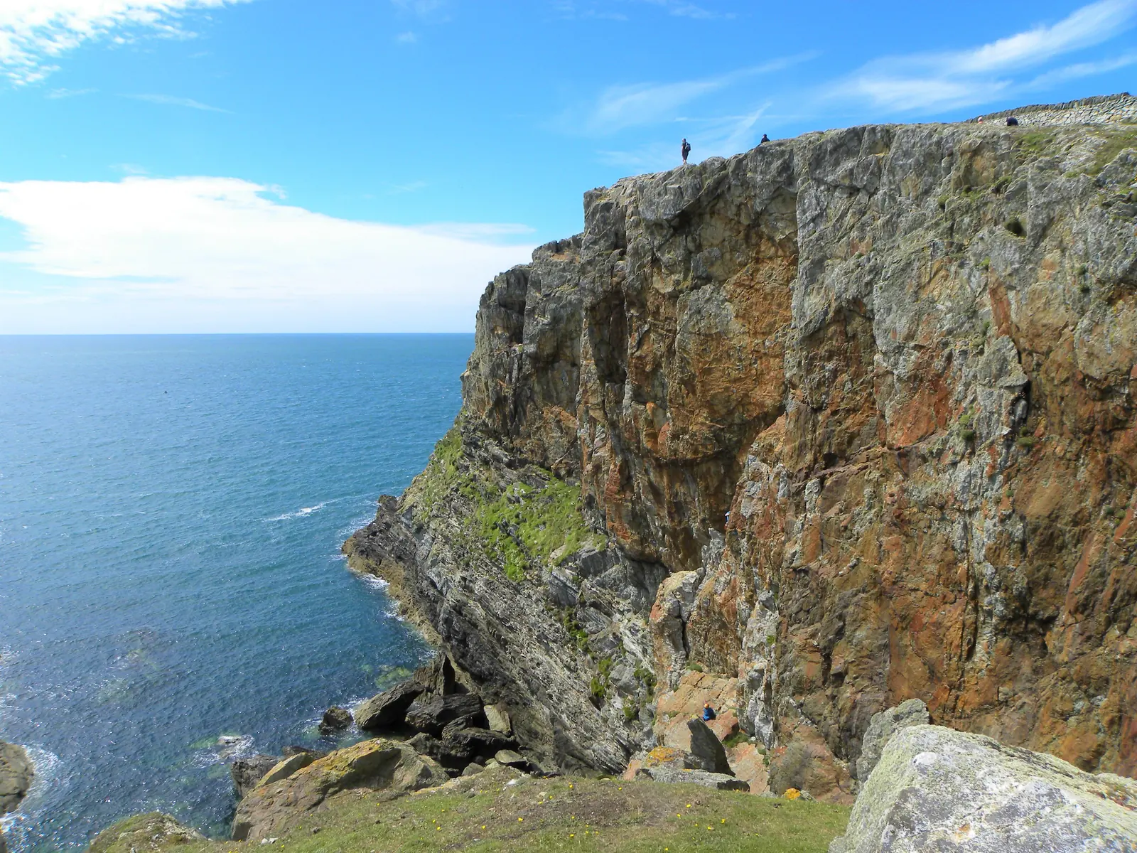 The cliffs at Porth Gwalch on the Rhoscolyn headland, with two small figures standing on the cliff top for scale.