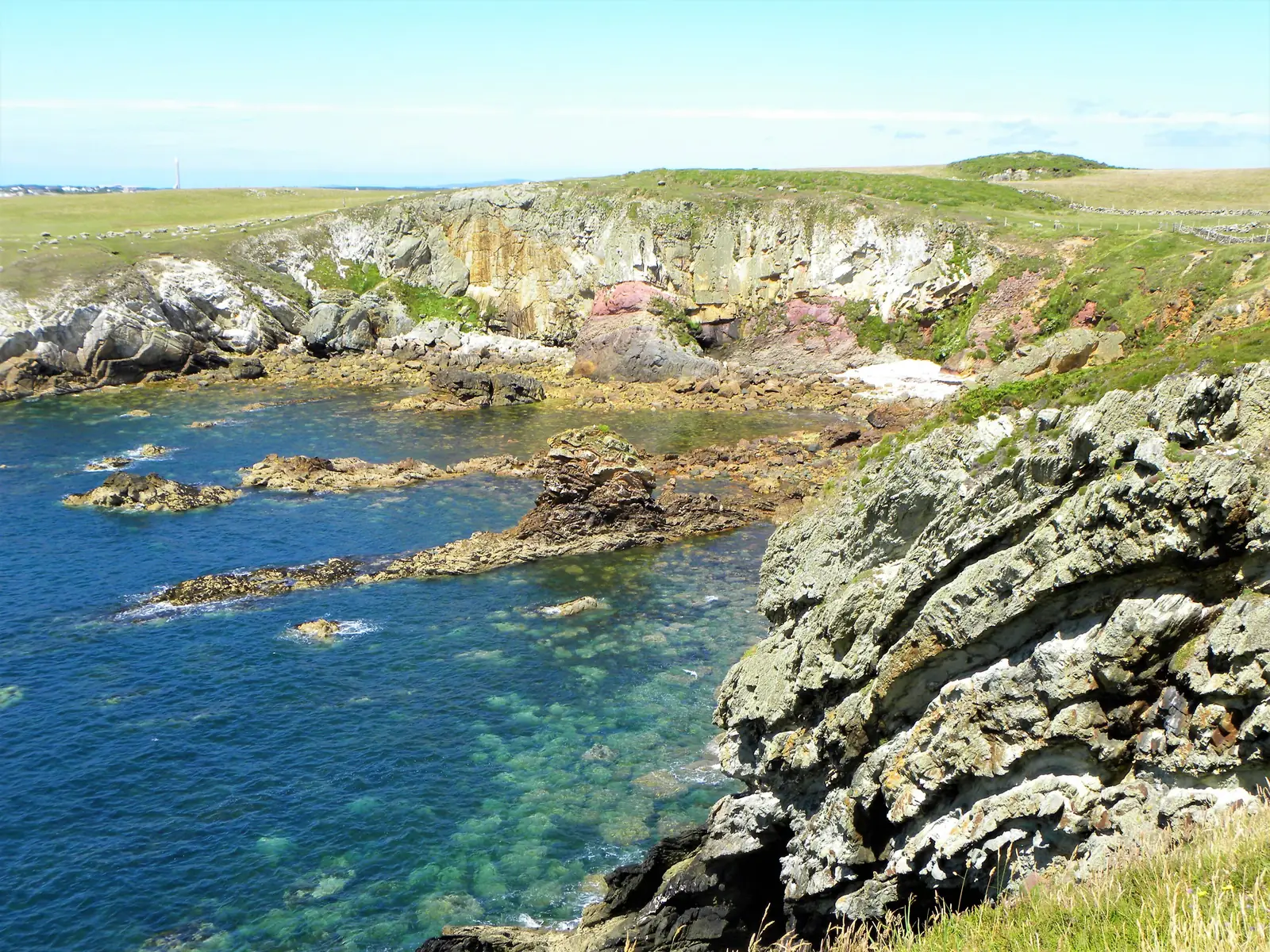 Turquoise water over a rocky cove at Porth Saint, Rhoscolyn, with banded and colour-shot quartzite in the cliffs above.