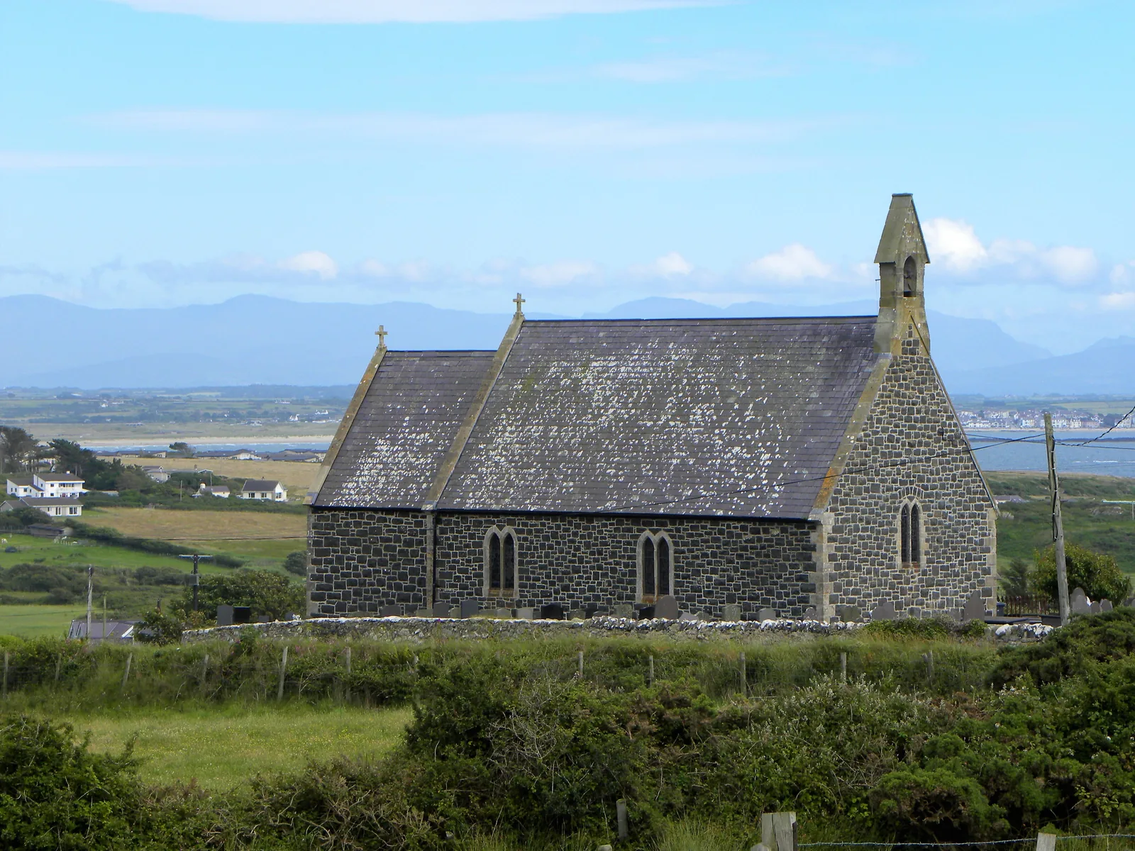 St Gwenfaen's Church at Rhoscolyn, a stone church with a bellcote, the mountains of Eryri on the skyline across the water behind it.