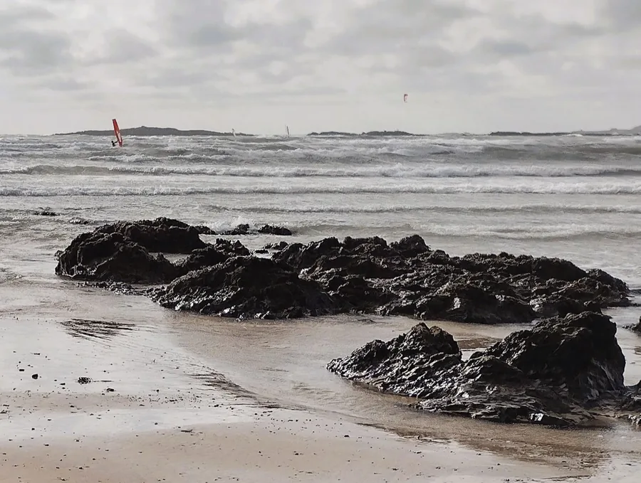 Waves breaking over dark rock at Traeth Crigyll, Rhosneigr, with windsurfers out on the water.
