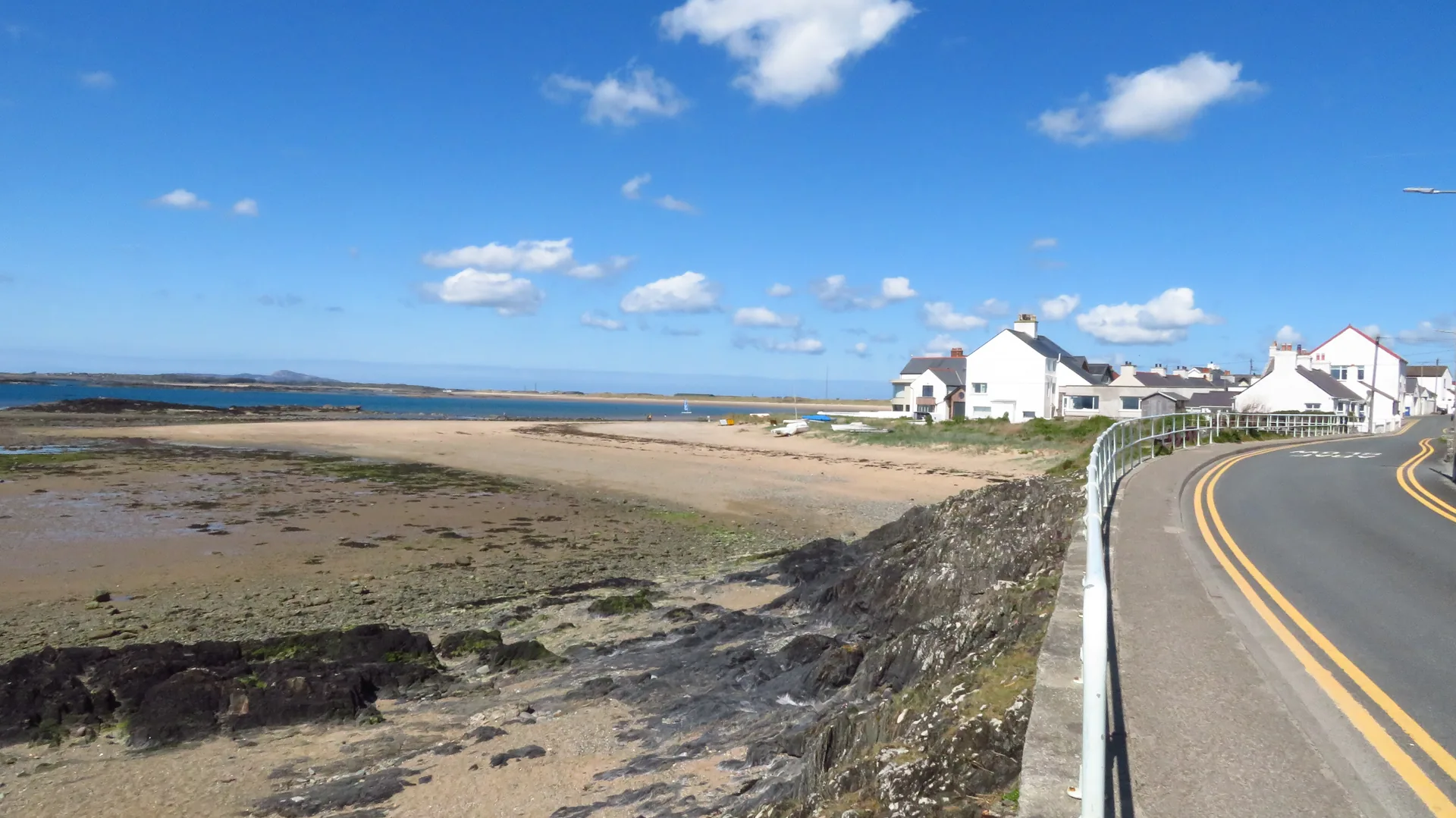 Rhosneigr from the coast road above the beach: wide pale sand curving away below white houses under a deep blue sky