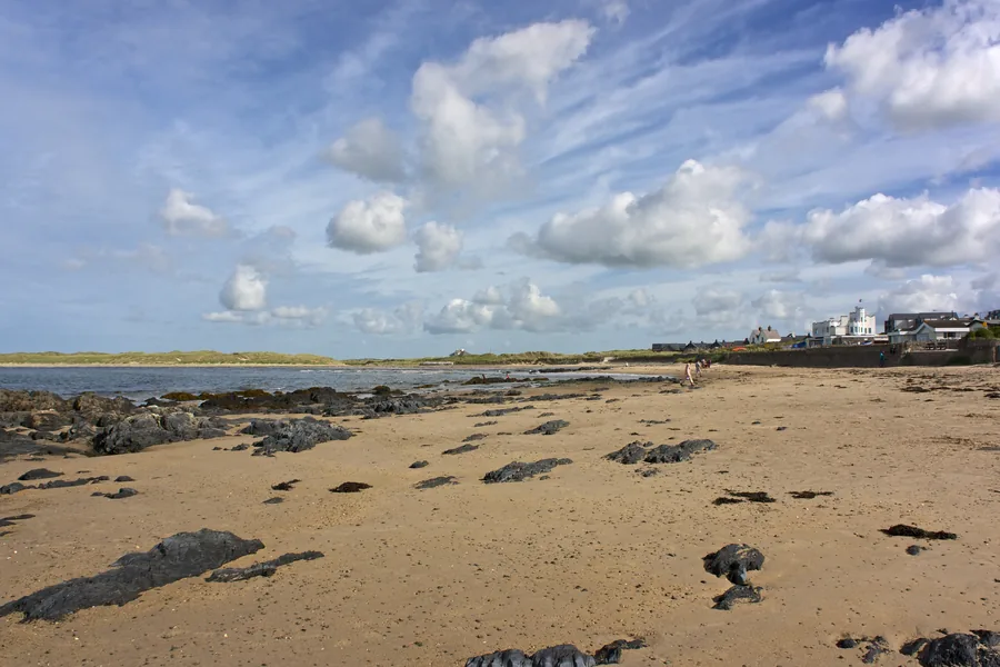 Rhosneigr beach at low water, wet sand and dark rock under a wide cloudscape, the village low on the skyline.