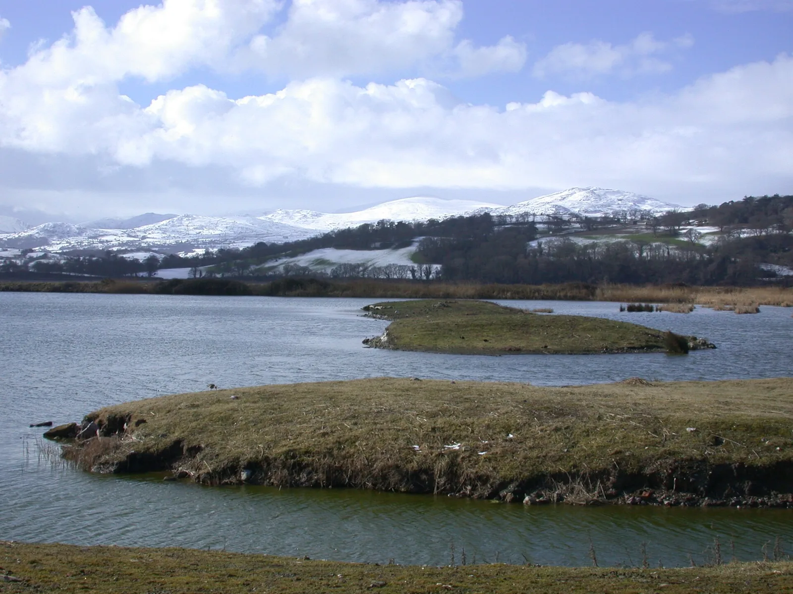 Grassy islands sitting in the open water of a lagoon at RSPB Conwy, with the snow-covered Carneddau mountains rising behind under a bright cloudy sky.