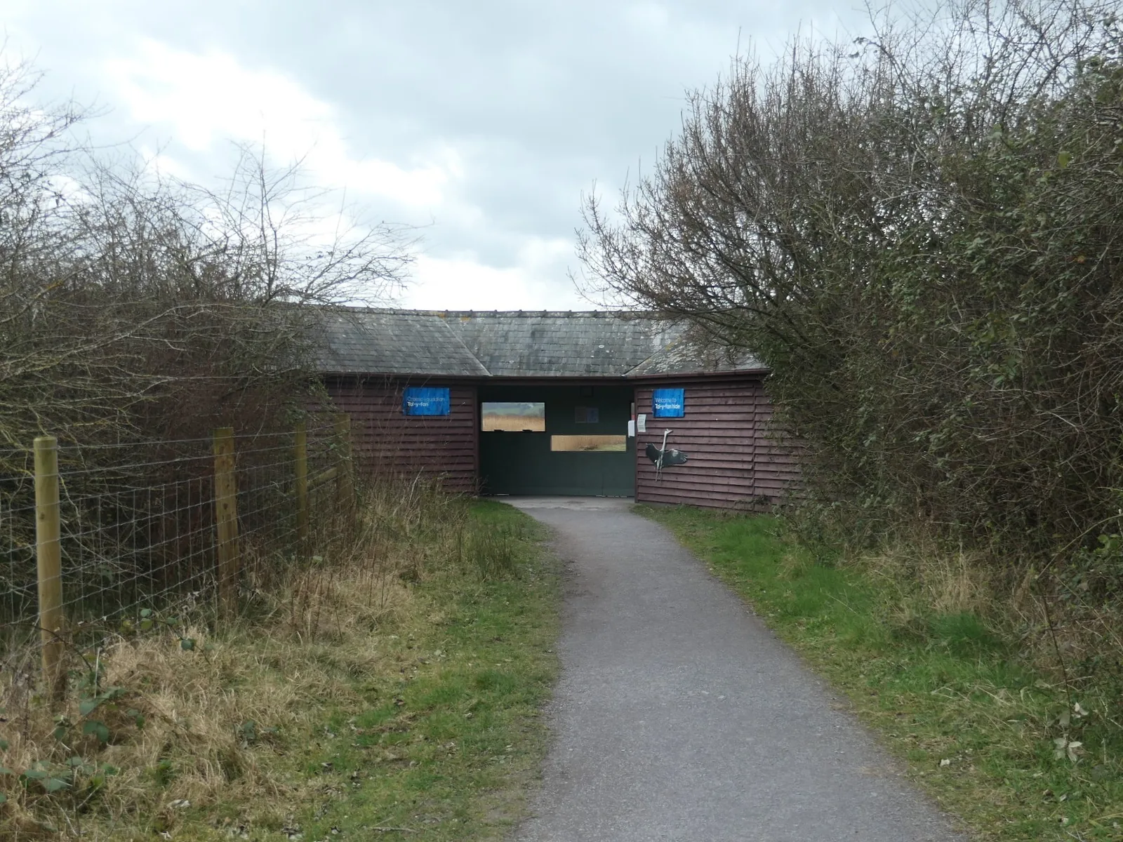 A low timber bird hide with a slate roof at the end of a gravel path at RSPB Conwy, flanked by bare hedgerow.