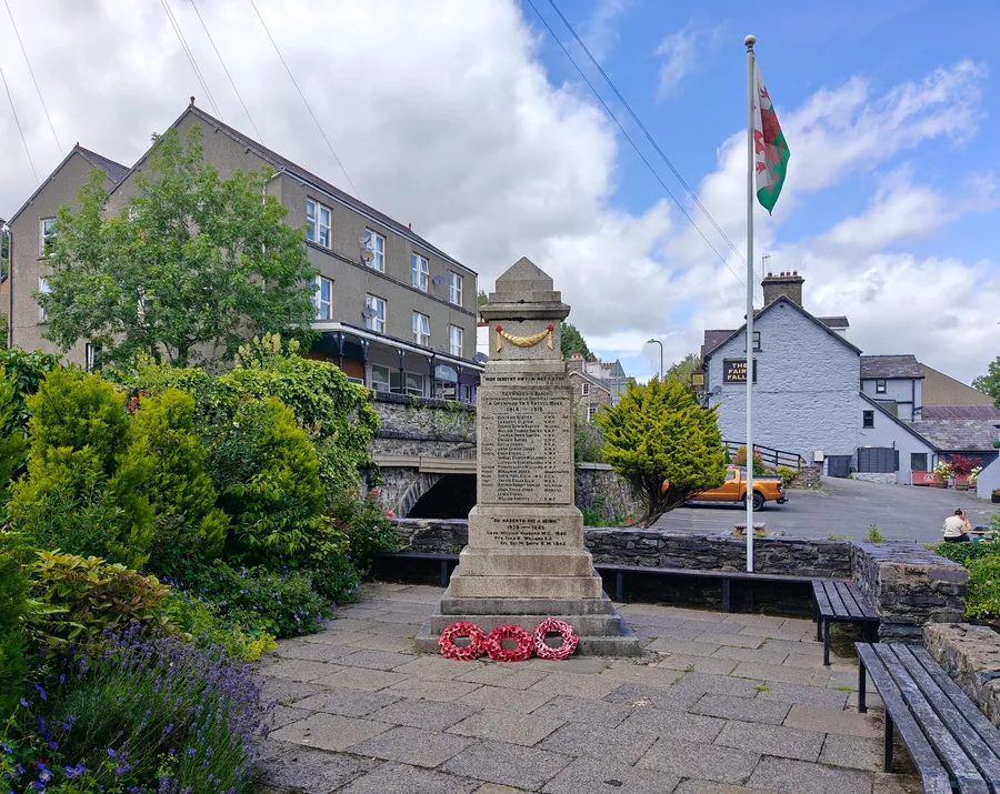 The war memorial at Trefriw with the Welsh flag flying beside it, the village and its wooded hillside behind.