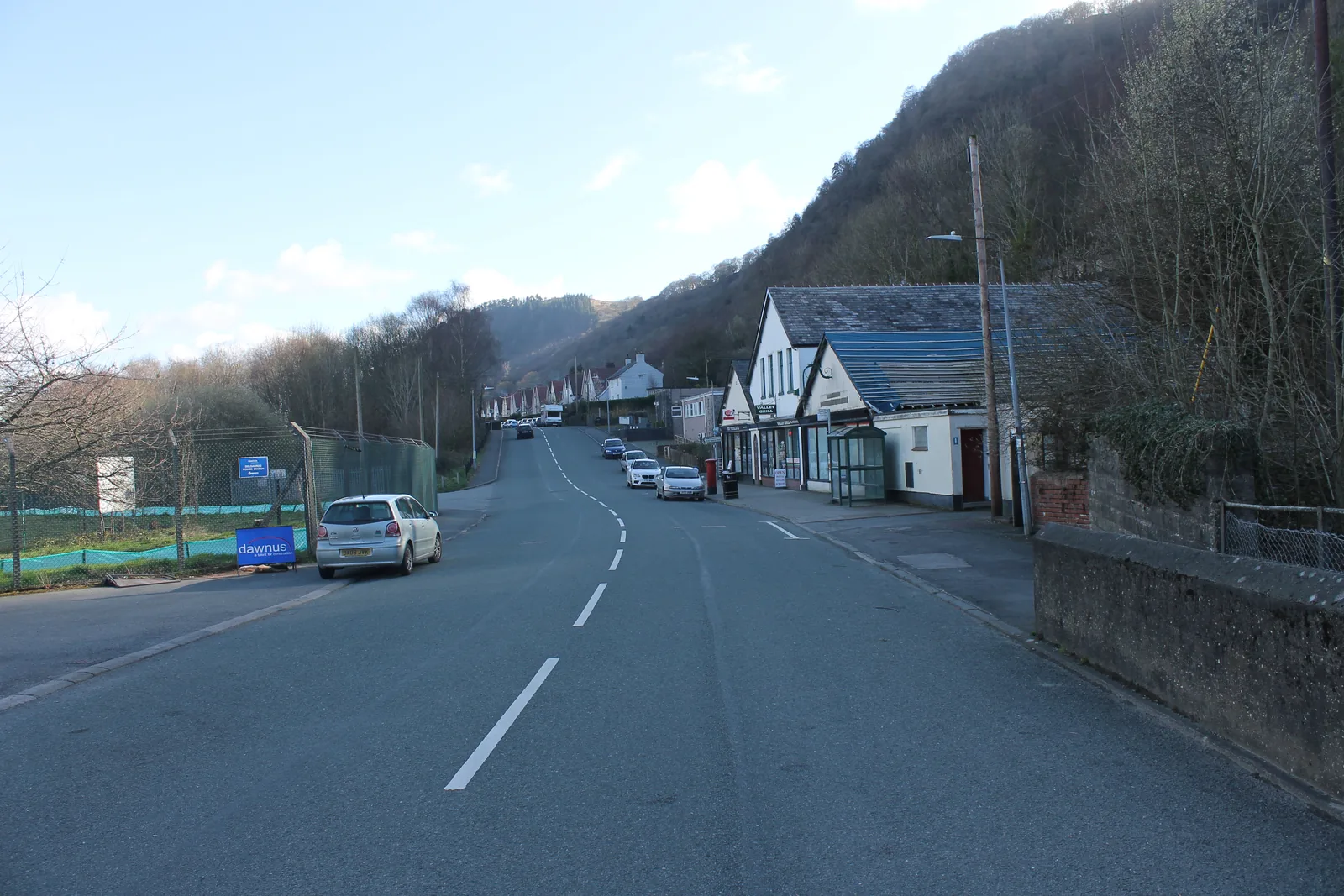 Shops and a bus shelter on the main road through Trefriw, the wooded slope rising immediately behind