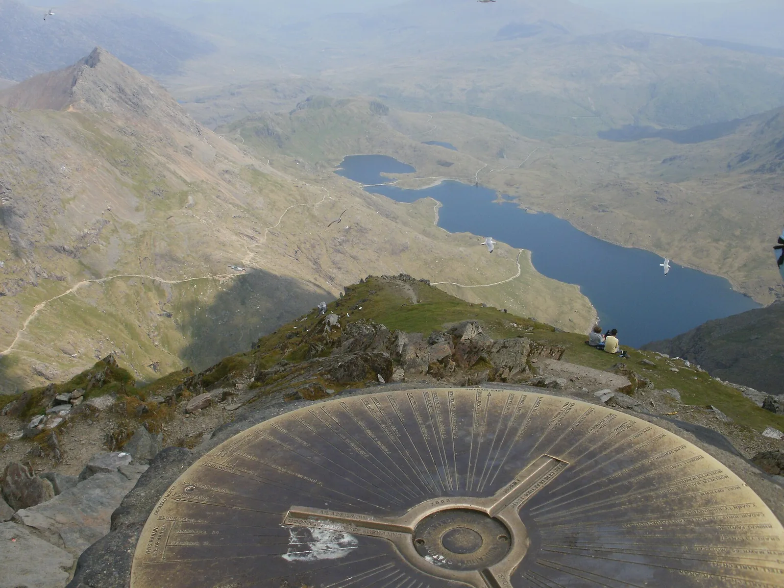 The view from the summit of Yr Wyddfa: a cast-bronze orientation table in the foreground, a long lake in the cwm below, a path zig-zagging down the slope, and a pinnacled ridge on the skyline