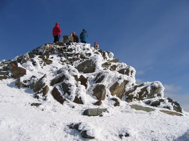 Walkers on the snow-covered summit cairn of Yr Wyddfa under a clear sky